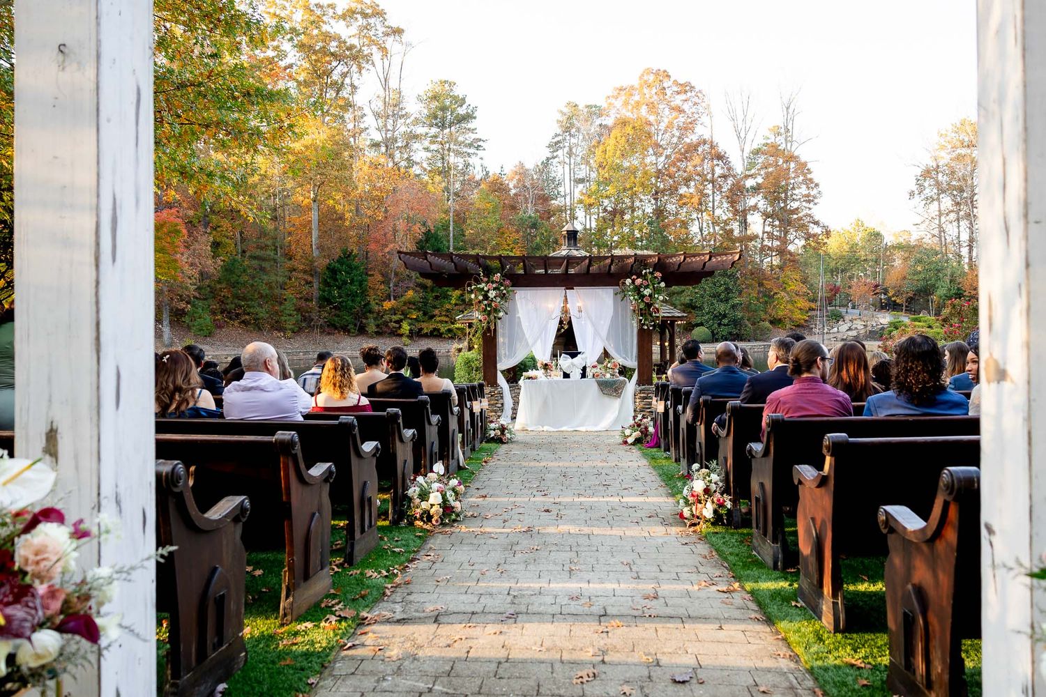 Outdoor fall wedding ceremony with guests seated on wooden pews along a stone aisle leading to a floral arch altar.