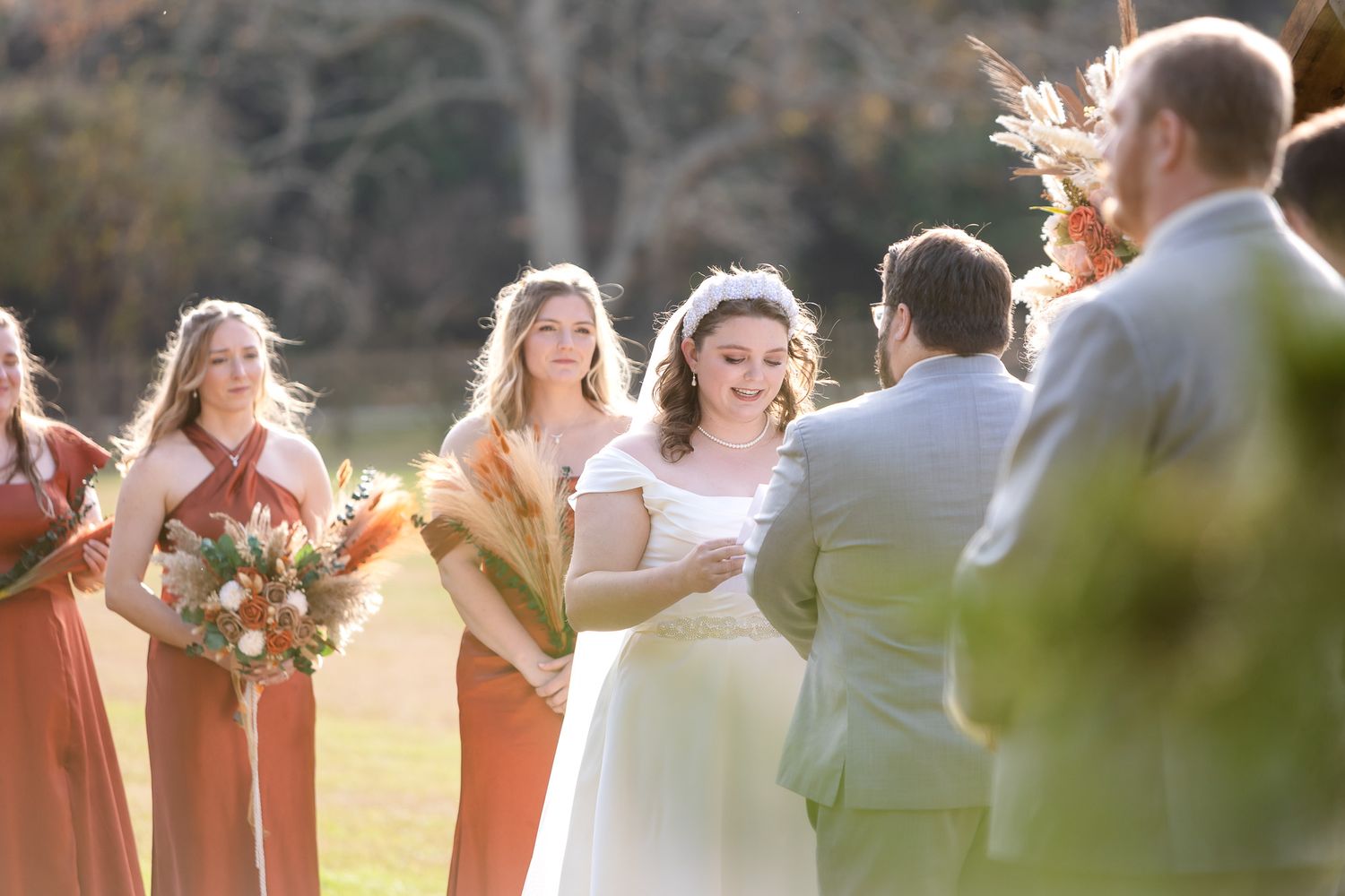 Bride smiling at groom during outdoor fall wedding ceremony with bridesmaids in rust orange dresses holding bouquets.