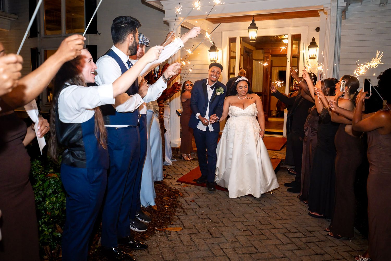 Bride and groom exit wedding venue through sparkler send-off tunnel with guests cheering at night.