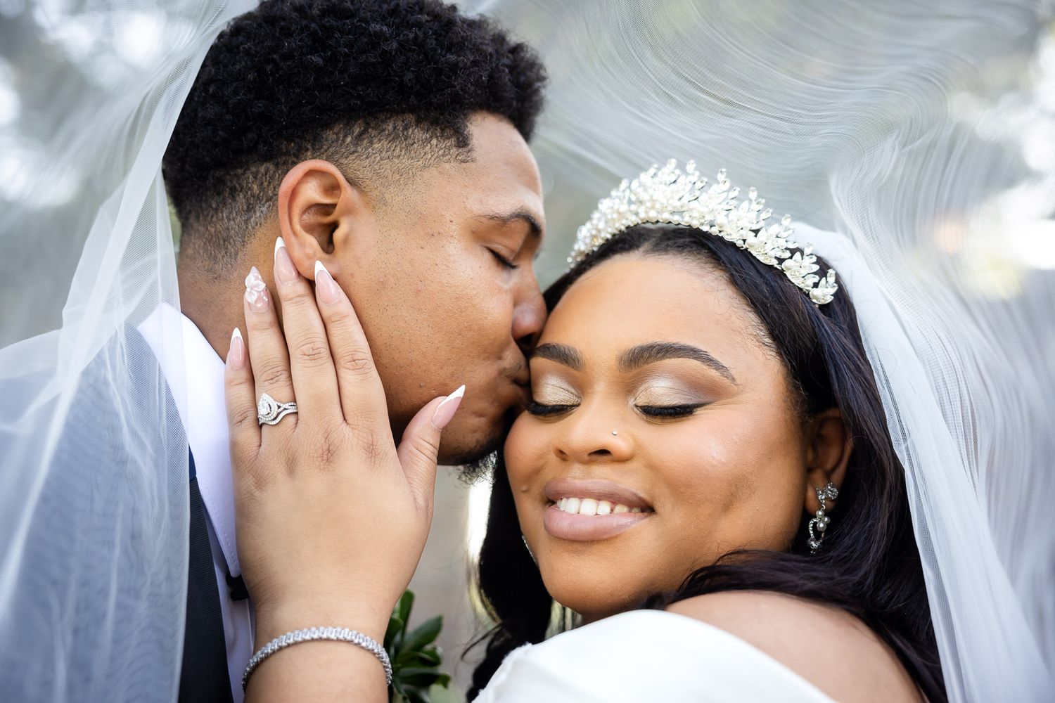 A groom kisses his bride on the cheek as she smiles, wearing a tiara and veil on their wedding day.