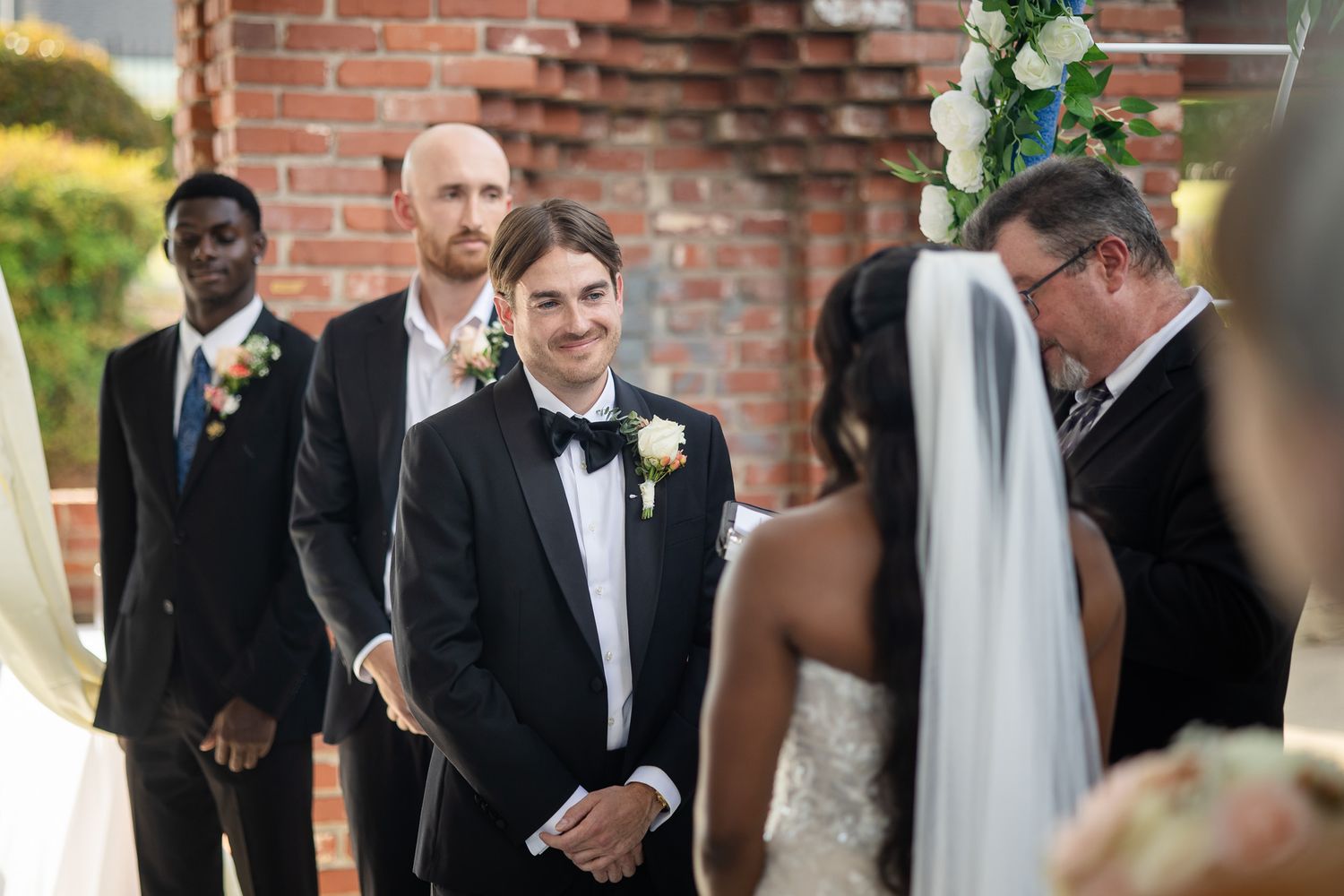 Groom smiling at bride during outdoor wedding ceremony with groomsmen and officiant at brick venue.