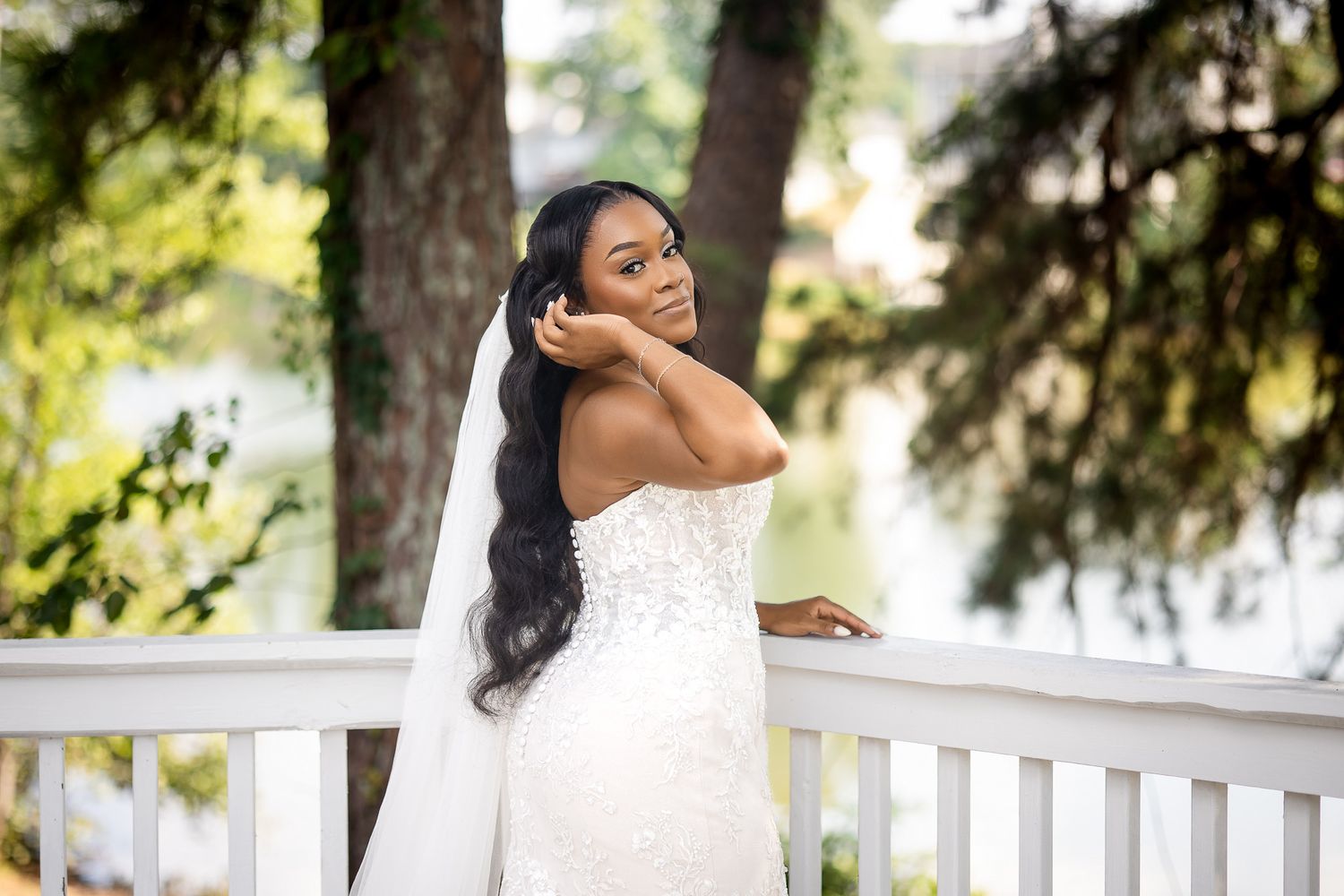 Elegant bride in a strapless lace wedding gown and veil poses gracefully on a white porch surrounded by trees.