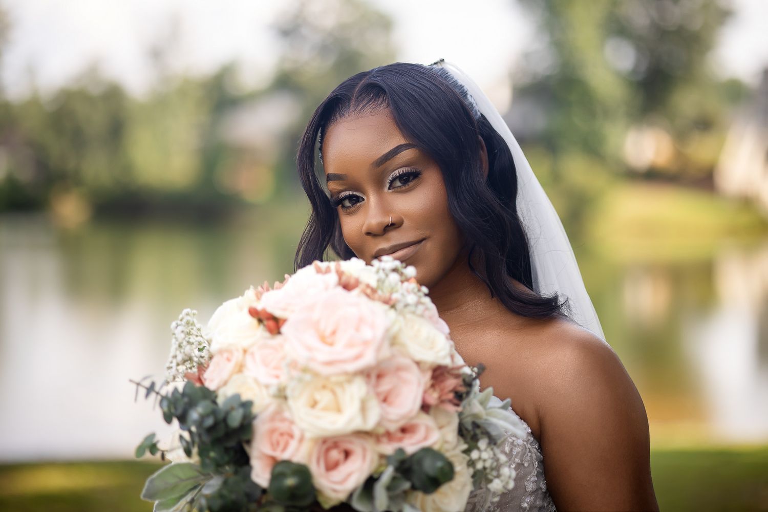 A beautiful Black bride in a white veil holds a blush pink bouquet outdoors near a serene pond backdrop.