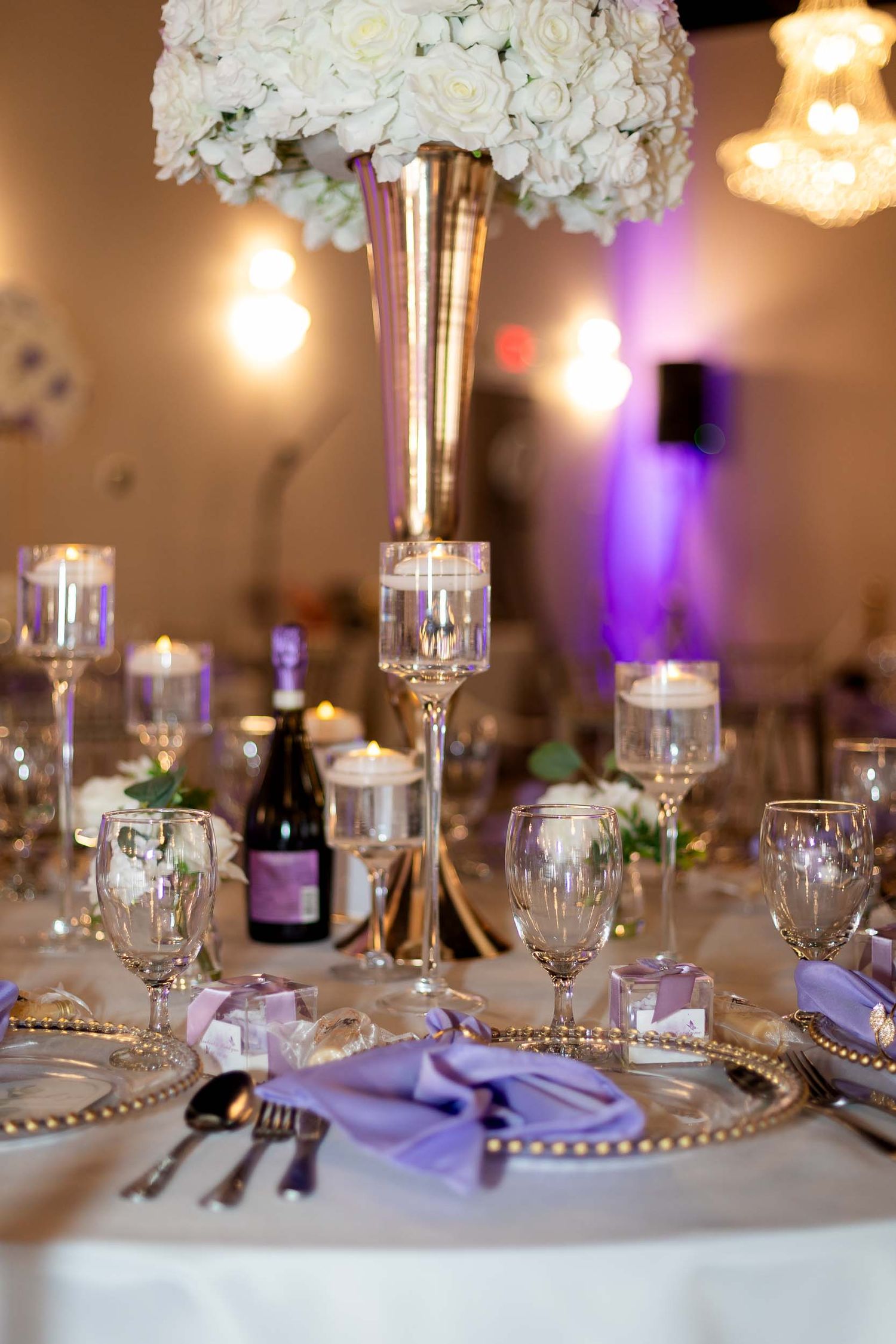 Elegant wedding reception table with lavender napkins, gold charger plates, crystal glasses, and tall white floral centerpiece.