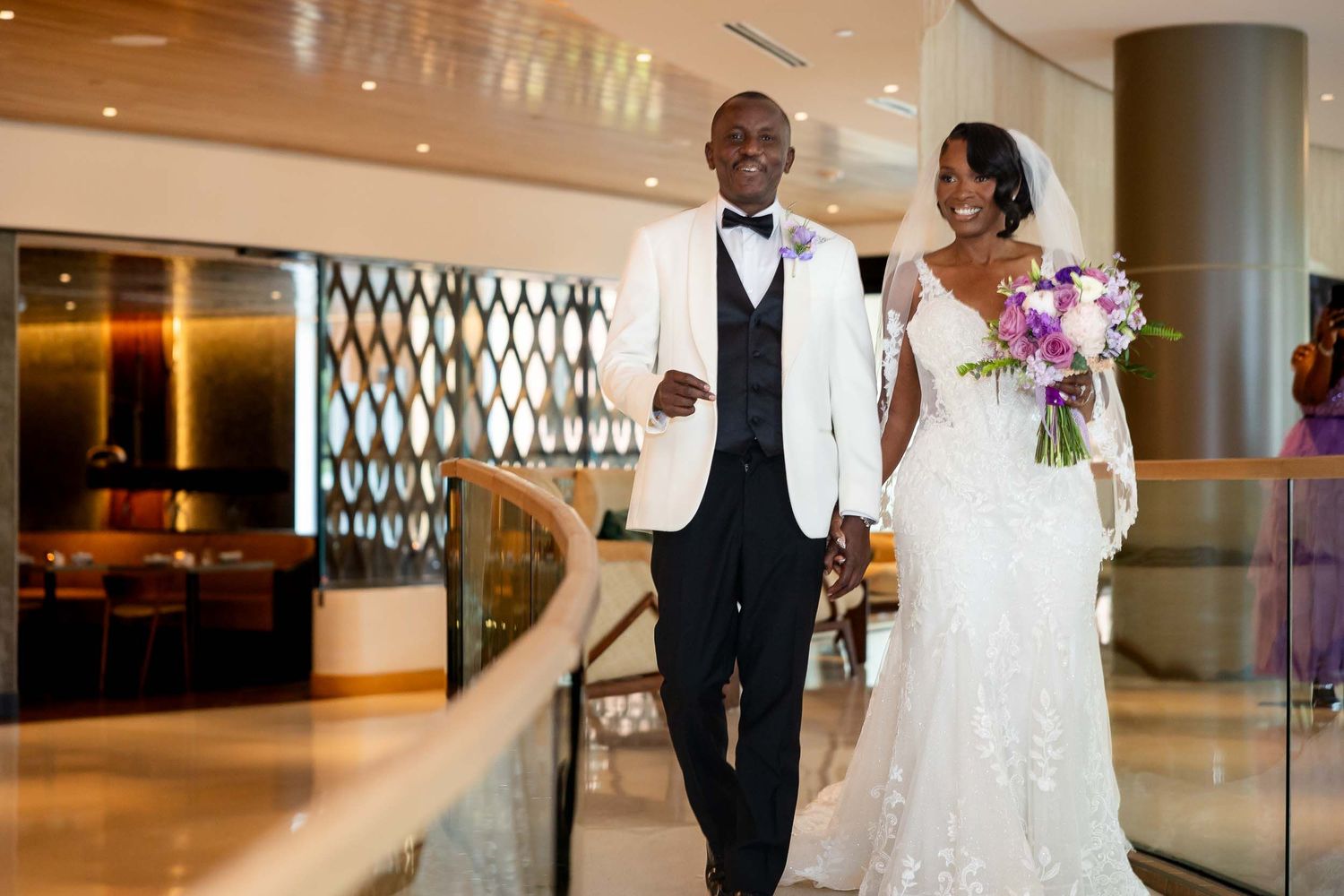 Elegant Black couple in wedding attire walk together in a modern venue, bride holding purple bouquet.