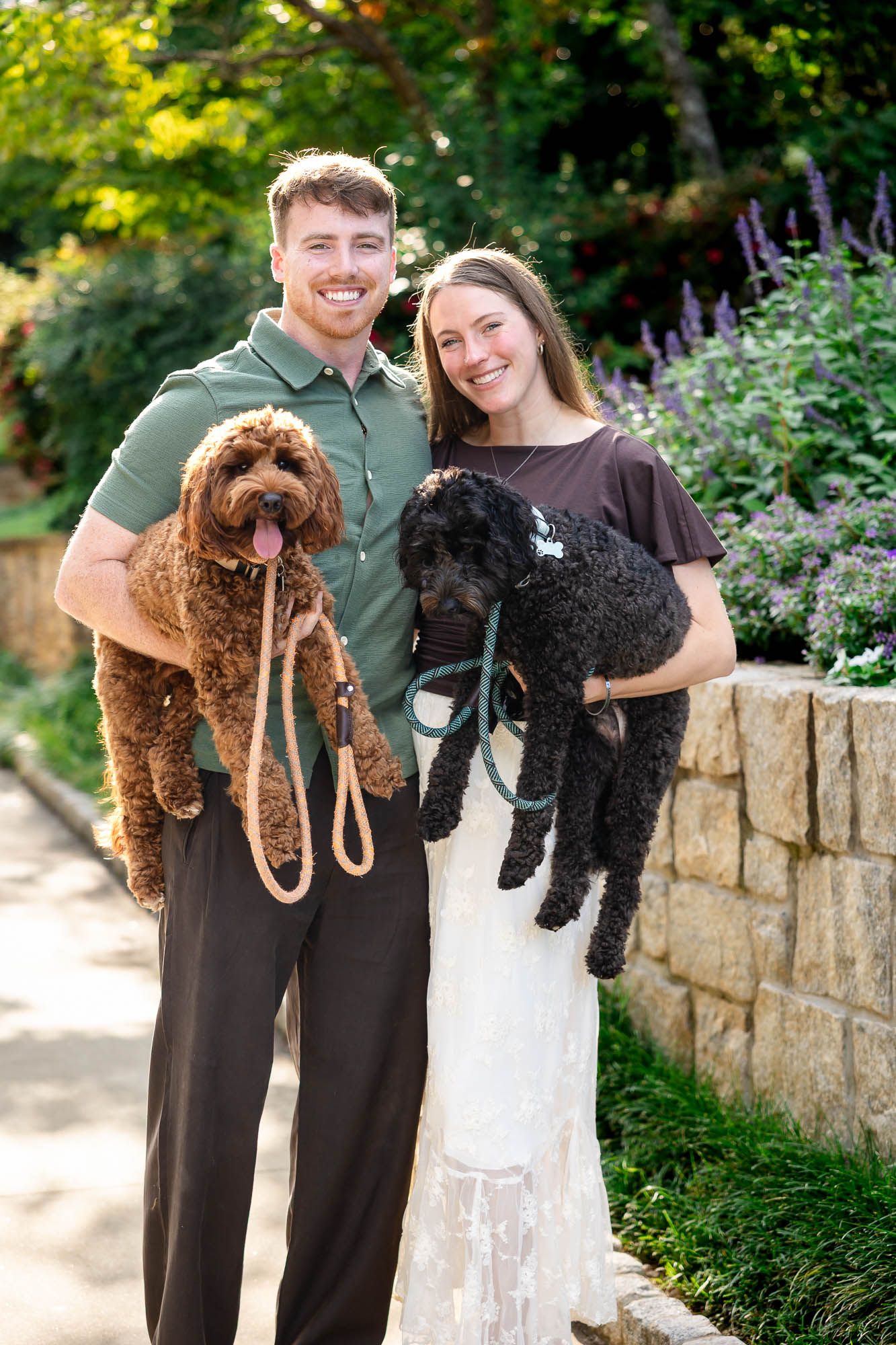 Smiling couple holding two fluffy dogs, a brown and black poodle mix, in a lush garden setting.