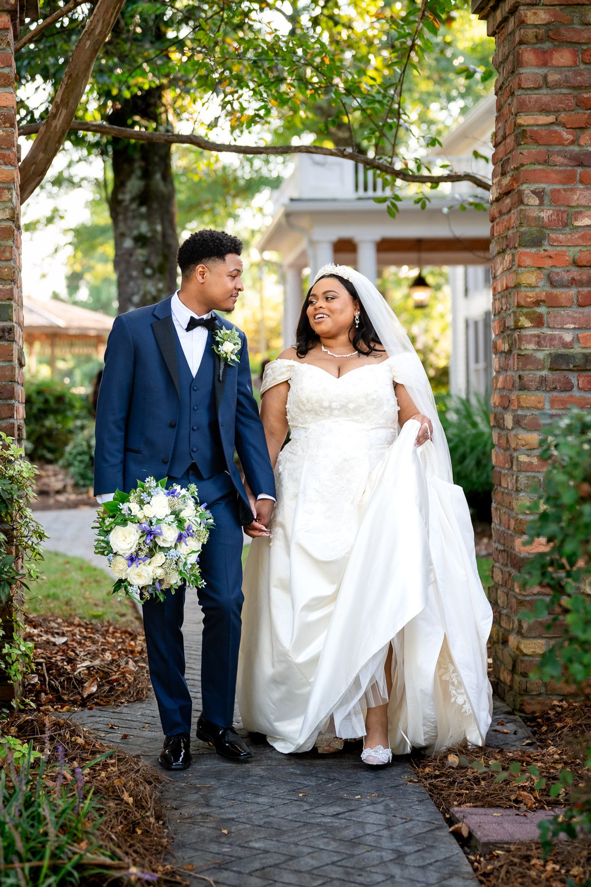 A bride and groom walk hand-in-hand outdoors, she in a white gown, he in a navy suit holding a floral bouquet.