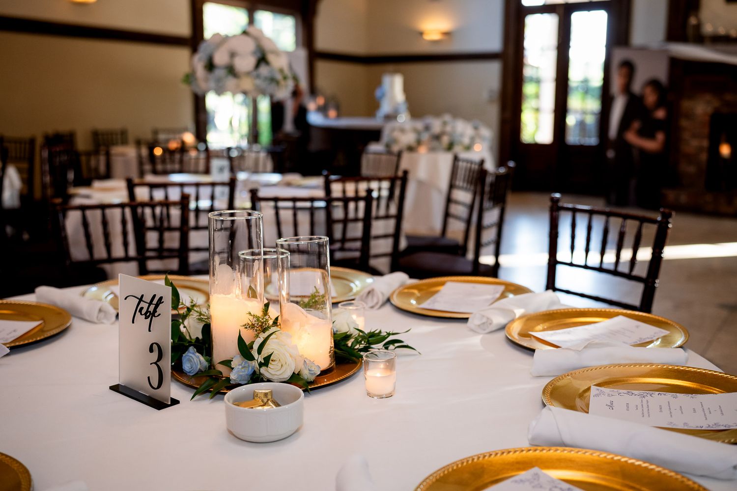Elegant wedding reception table with gold charger plates, white candles, floral centerpiece, and table number card in a ballroom.