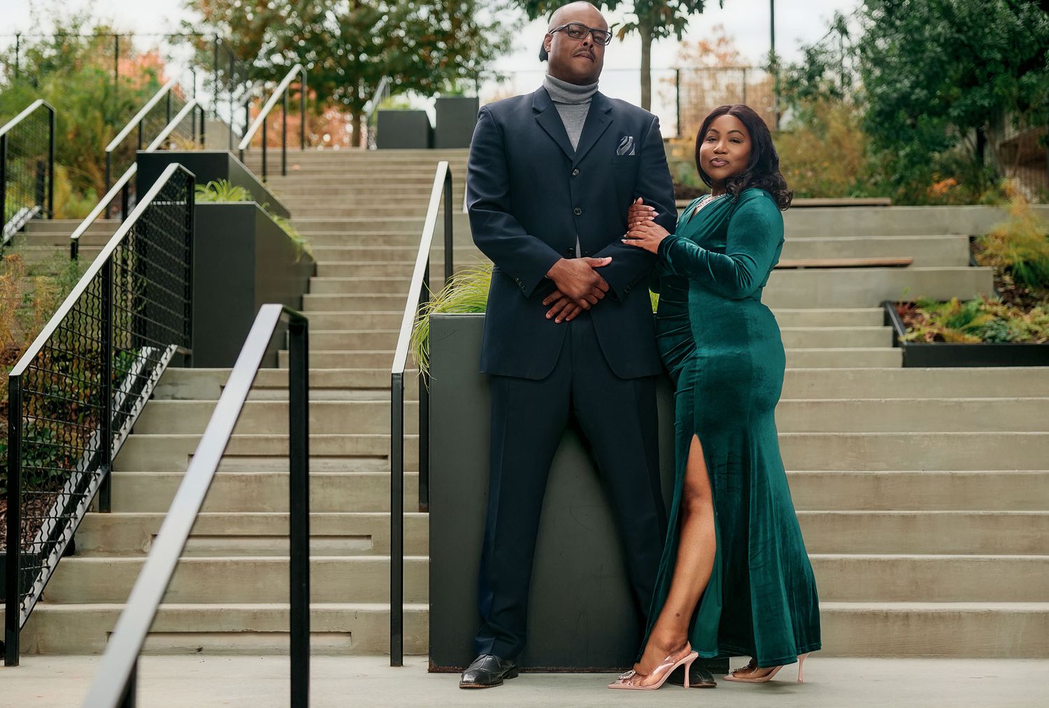 Elegant couple posing on steps, man in navy suit, woman in emerald green velvet gown, outdoor urban setting.