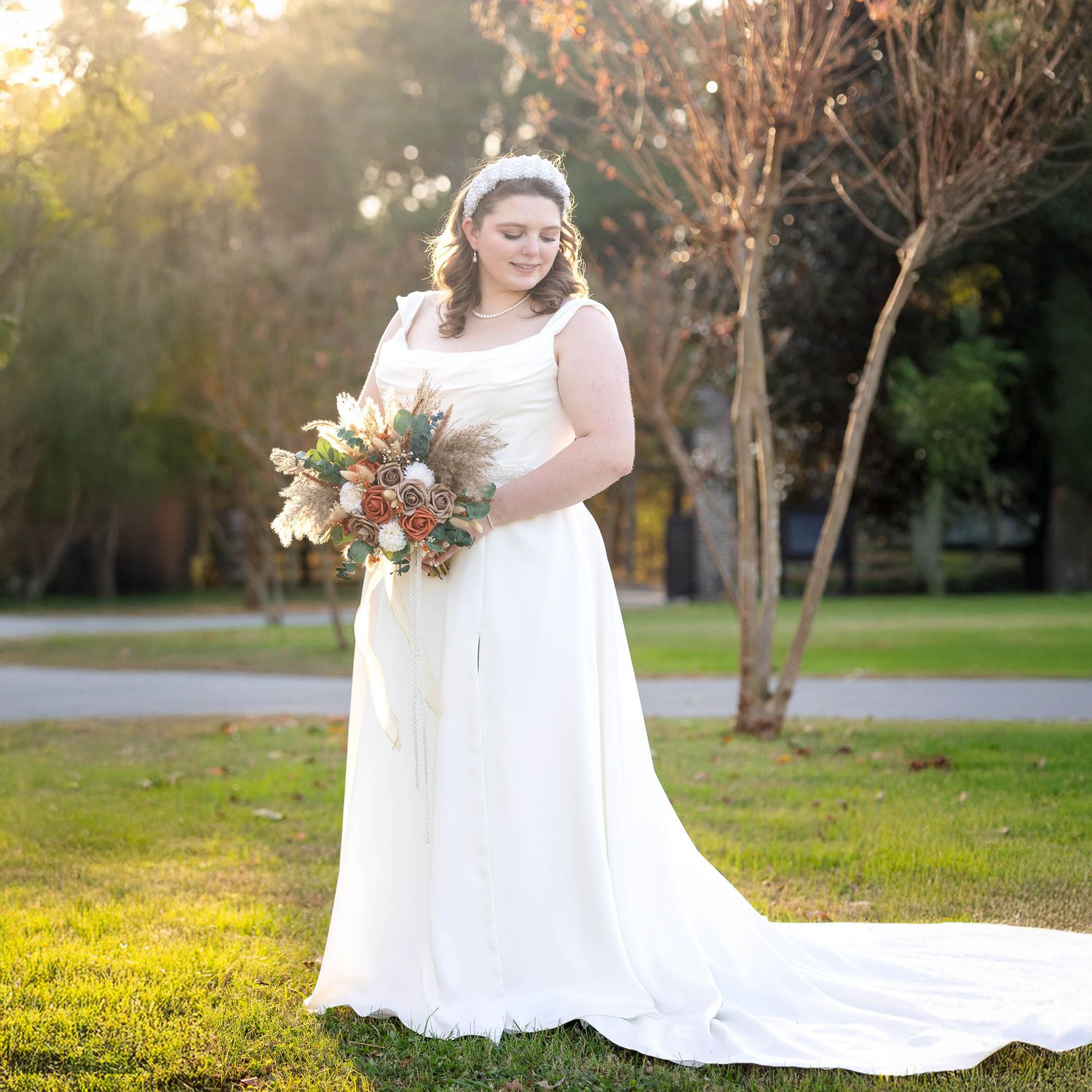 Bride in white A-line gown holding autumn bouquet stands on green lawn with golden sunlight and trees behind her.