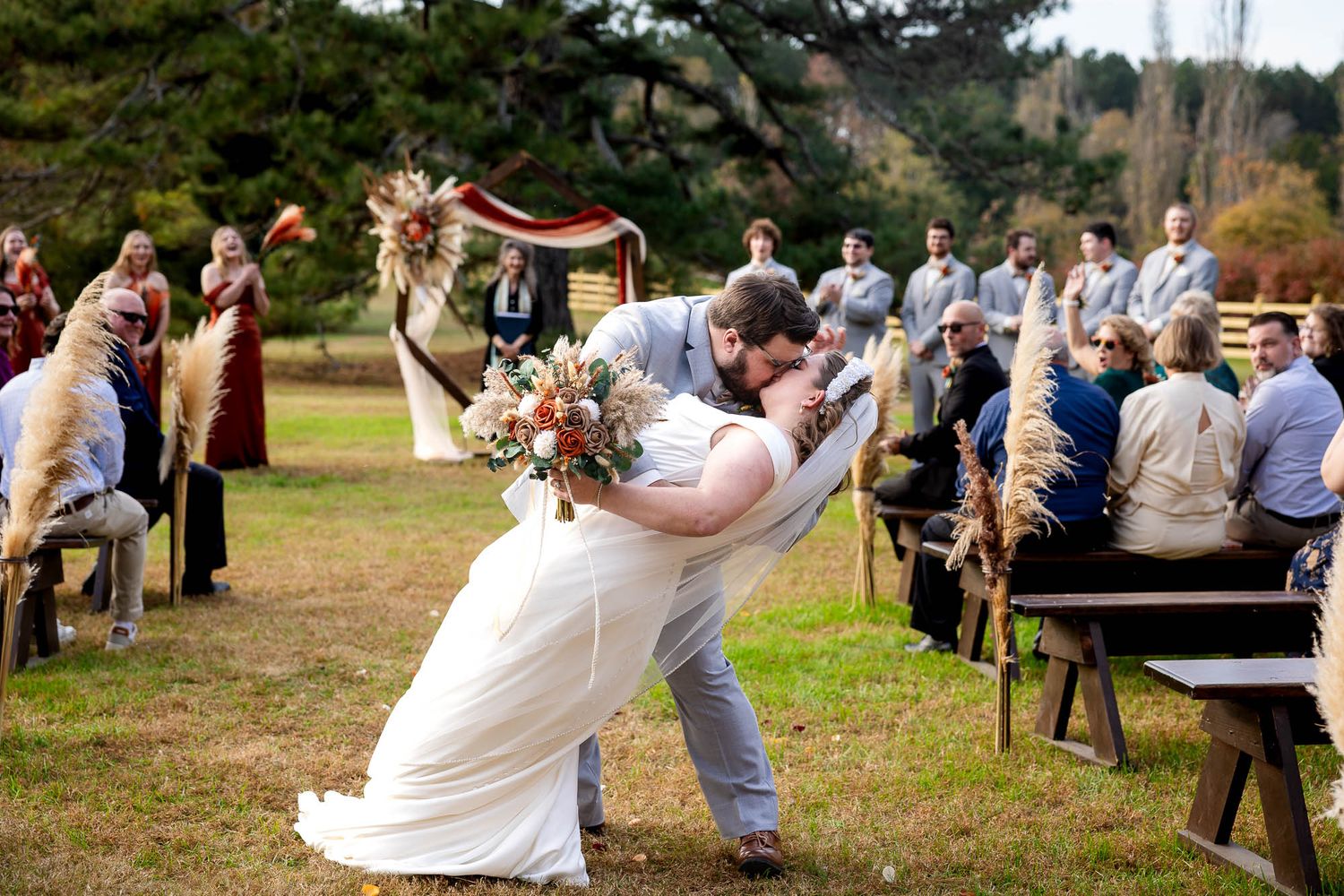 Groom dipping bride for romantic kiss at outdoor fall wedding ceremony with guests seated on wooden benches.