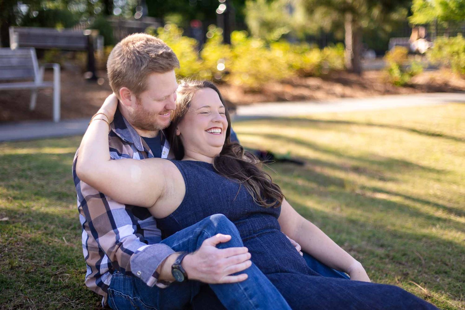 Happy couple sitting on grass in a sunny park, laughing and embracing in a candid engagement photo.