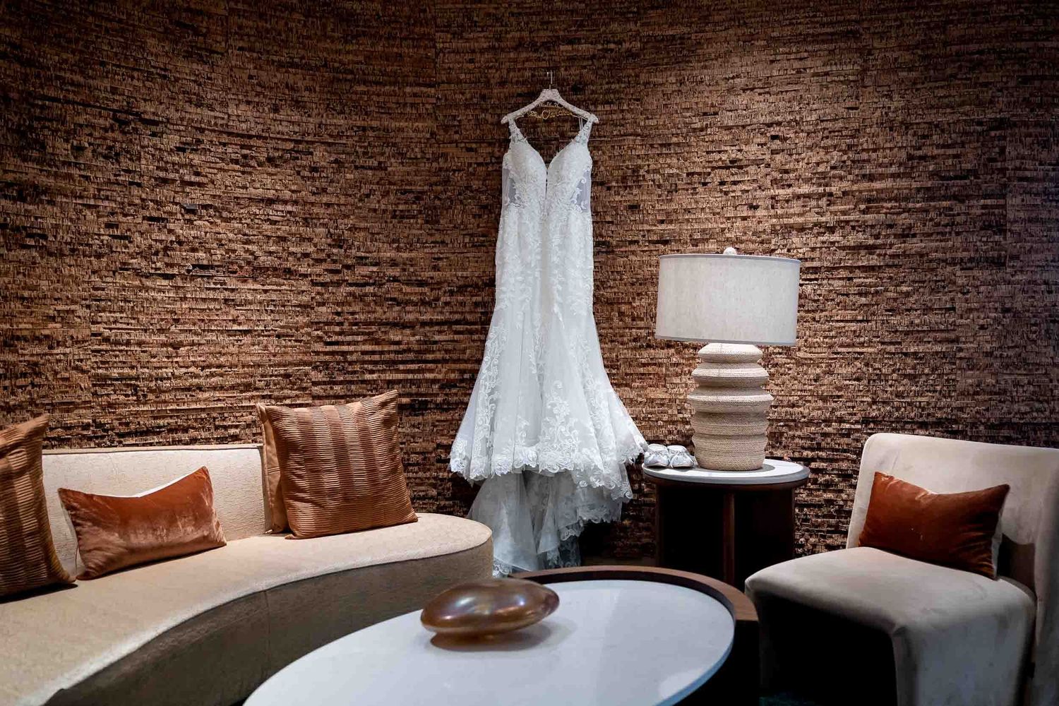 Elegant white lace wedding dress hanging on a brick wall in a warmly lit hotel suite with neutral furnishings.