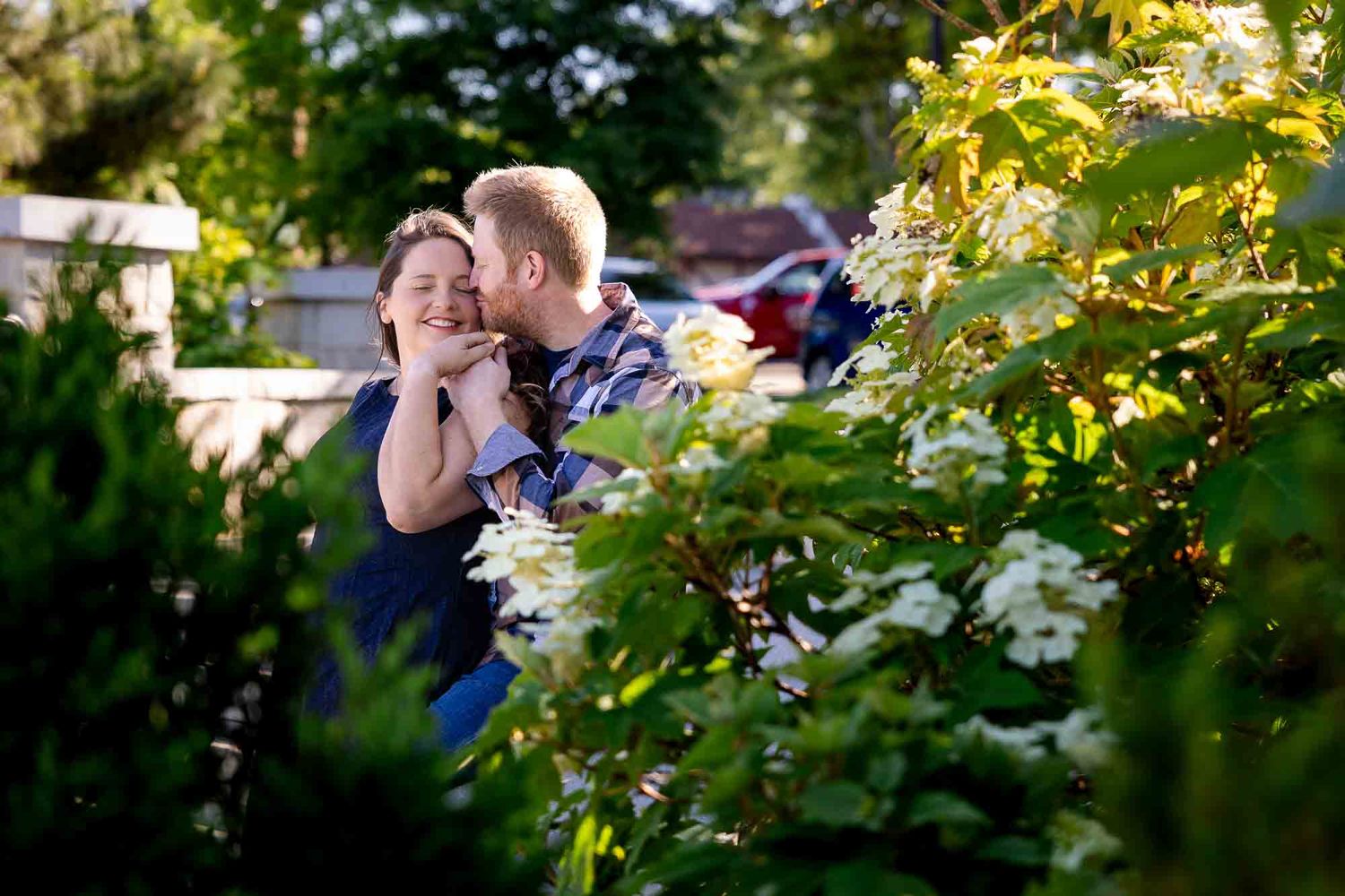 Couple sharing a romantic kiss among lush green bushes with white flowers in a sunny garden setting.