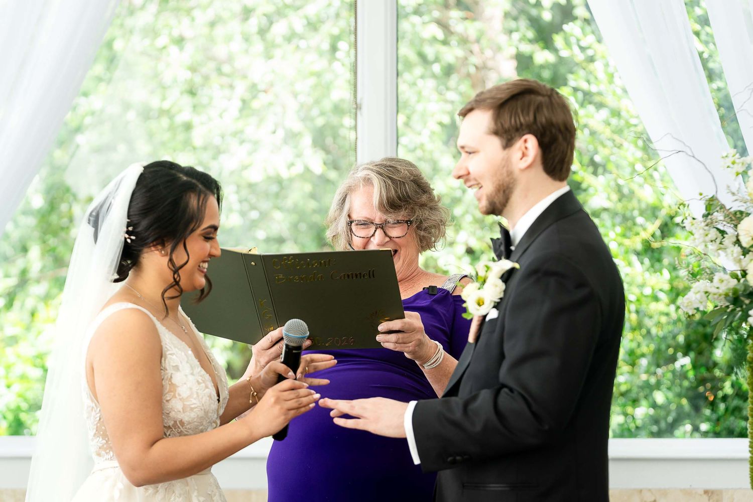 Bride and groom exchanging rings during wedding ceremony with officiant in purple dress holding folder.