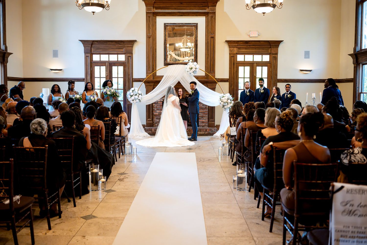 Elegant indoor wedding ceremony with bride and groom at floral arch altar, white aisle runner, and seated guests in wood-paneled venue.