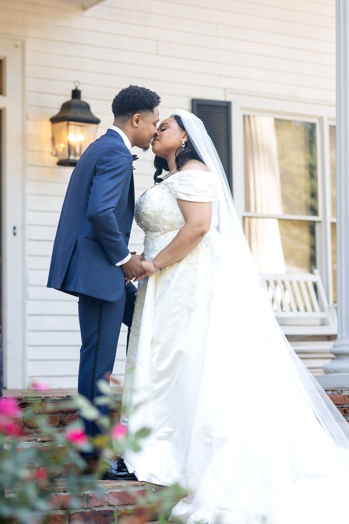 A newlywed couple shares a kiss on porch steps, bride in white gown with veil, groom in navy suit.