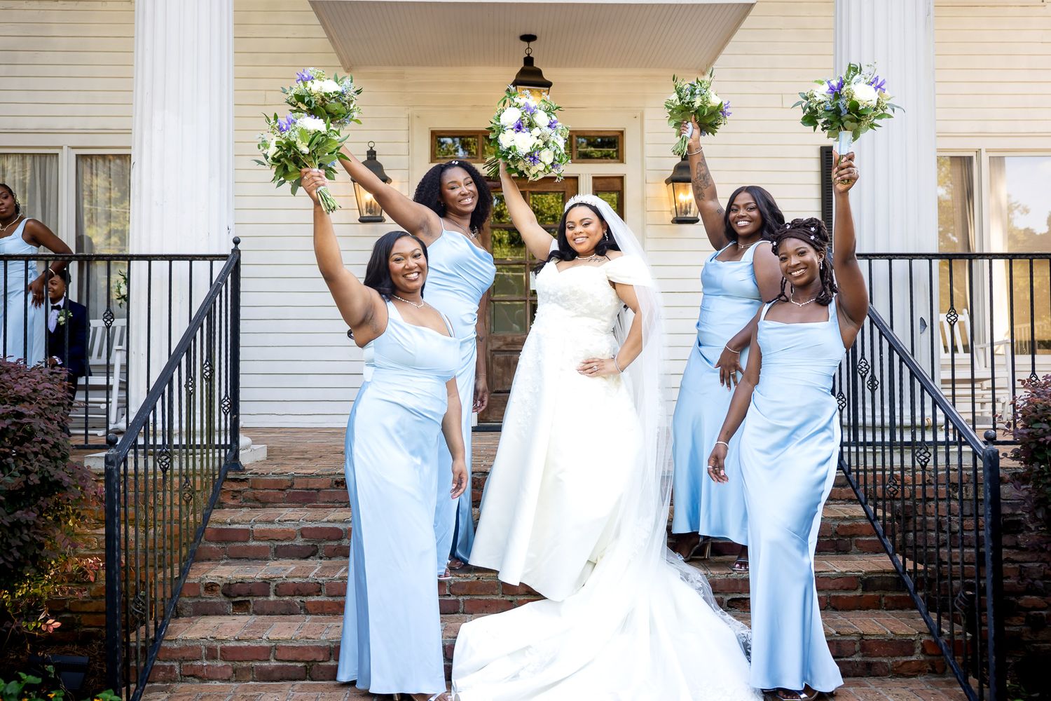 Bride in white gown celebrates with four bridesmaids in light blue dresses, holding bouquets on steps of elegant venue.