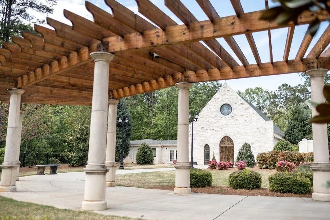 A wooden pergola with white columns provides shade over an outdoor patio area beside a white stone building.