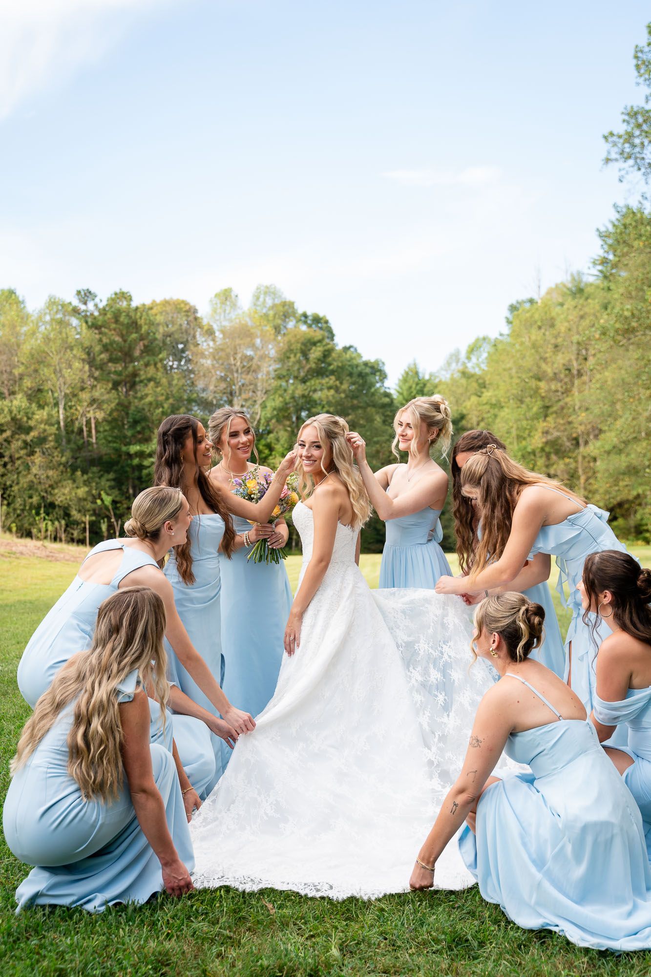 A group in light blue dresses sits together on grass, creating a joyful wedding party scene outdoors.