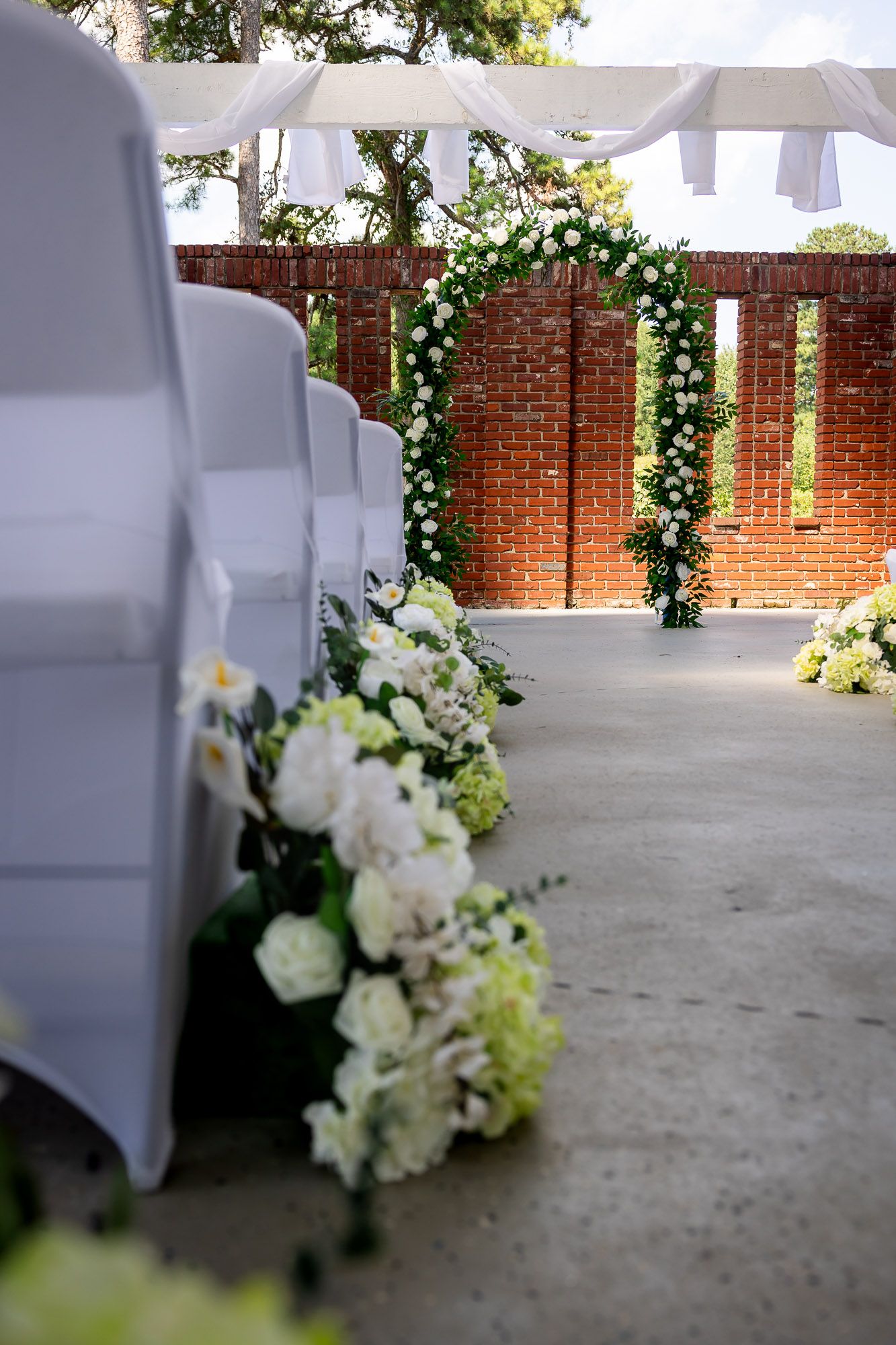 Elegant outdoor wedding ceremony setup with white floral arch, chair covers, and flower arrangements against a brick wall backdrop.