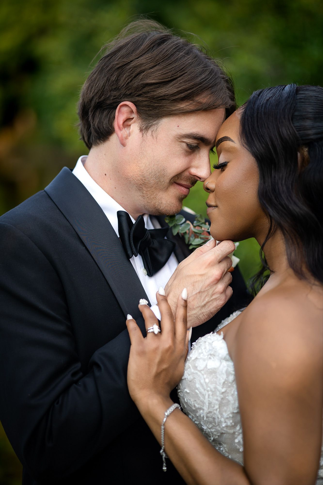 Interracial couple in wedding attire sharing an intimate moment, groom in tuxedo and bride in white lace gown outdoors.