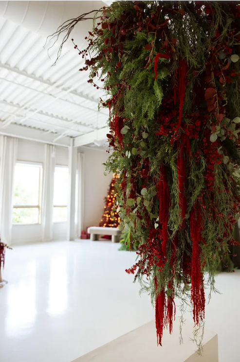Lush Christmas greenery arrangement with red berries and ribbons hanging from ceiling in bright white studio space.