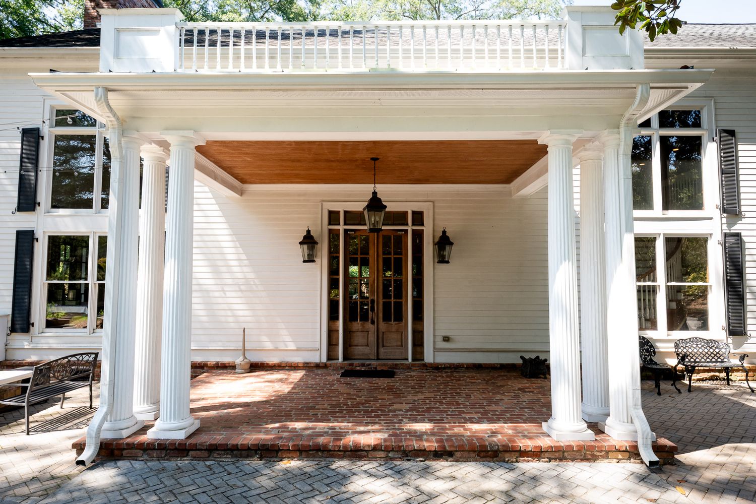 Elegant white colonial home entrance with brick patio, white columns, wood ceiling, and lantern sconces flanking a glass door.