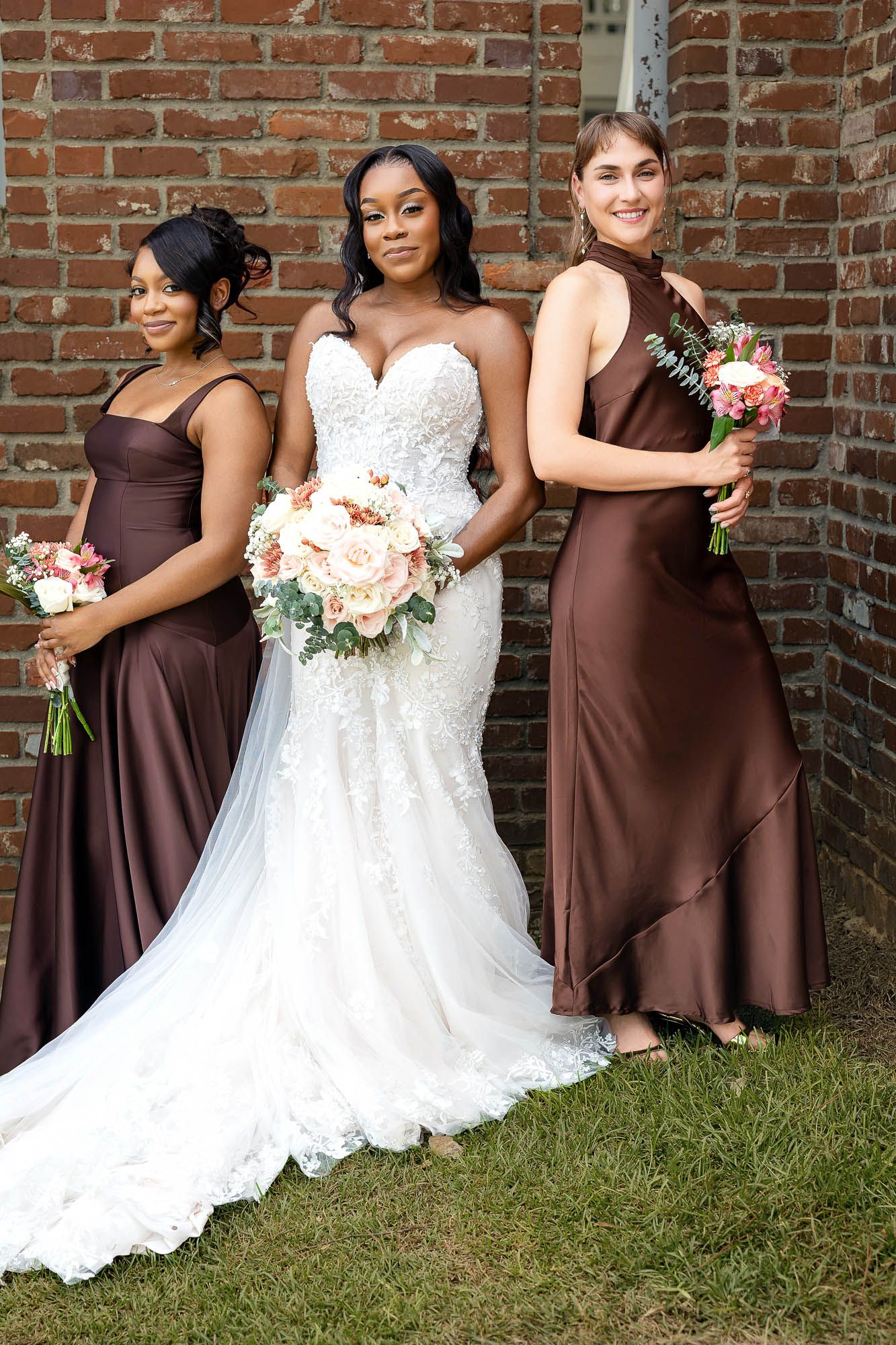 Bride in white lace gown poses with two bridesmaids in brown satin dresses holding bouquets.