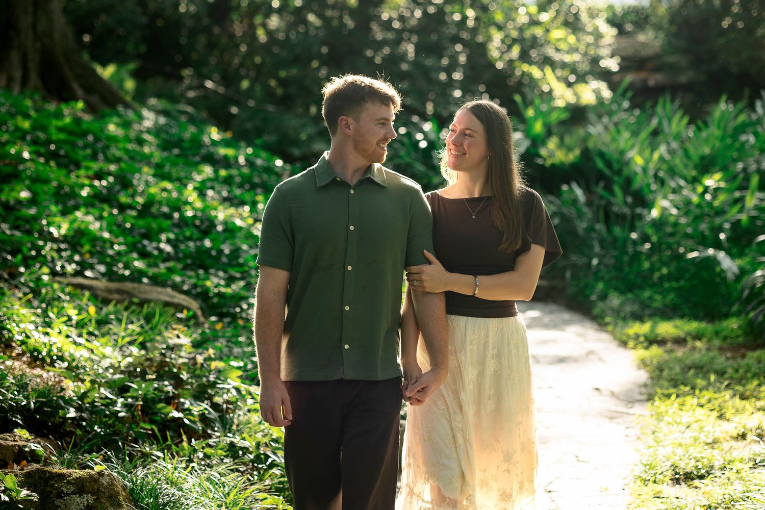 A happy couple walks hand-in-hand along a sunlit garden path surrounded by lush green foliage.