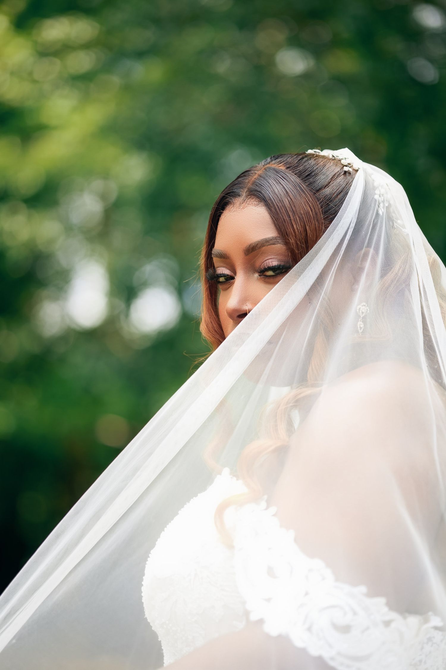 A bride in white gown stands outdoors wearing a delicate sheer veil against a blurred natural green background.