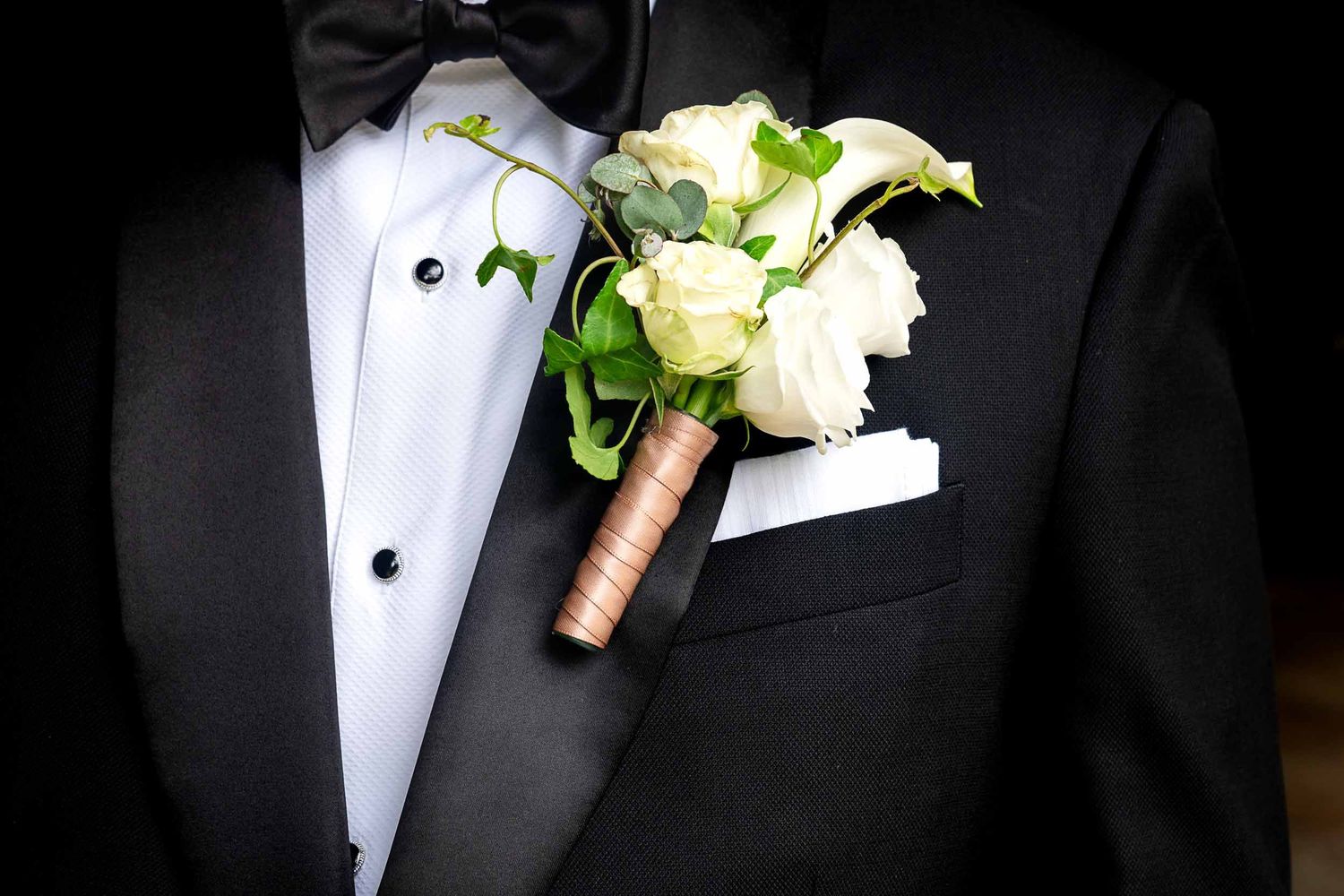 Groom in black tuxedo with white boutonniere featuring roses and greenery wrapped in gold ribbon on lapel.