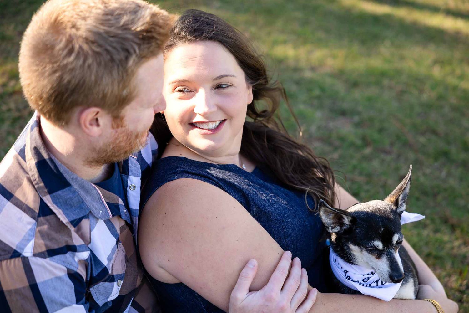 Smiling couple sitting outdoors on grass, woman holding small black and white dog wearing a bandana, man embracing her.