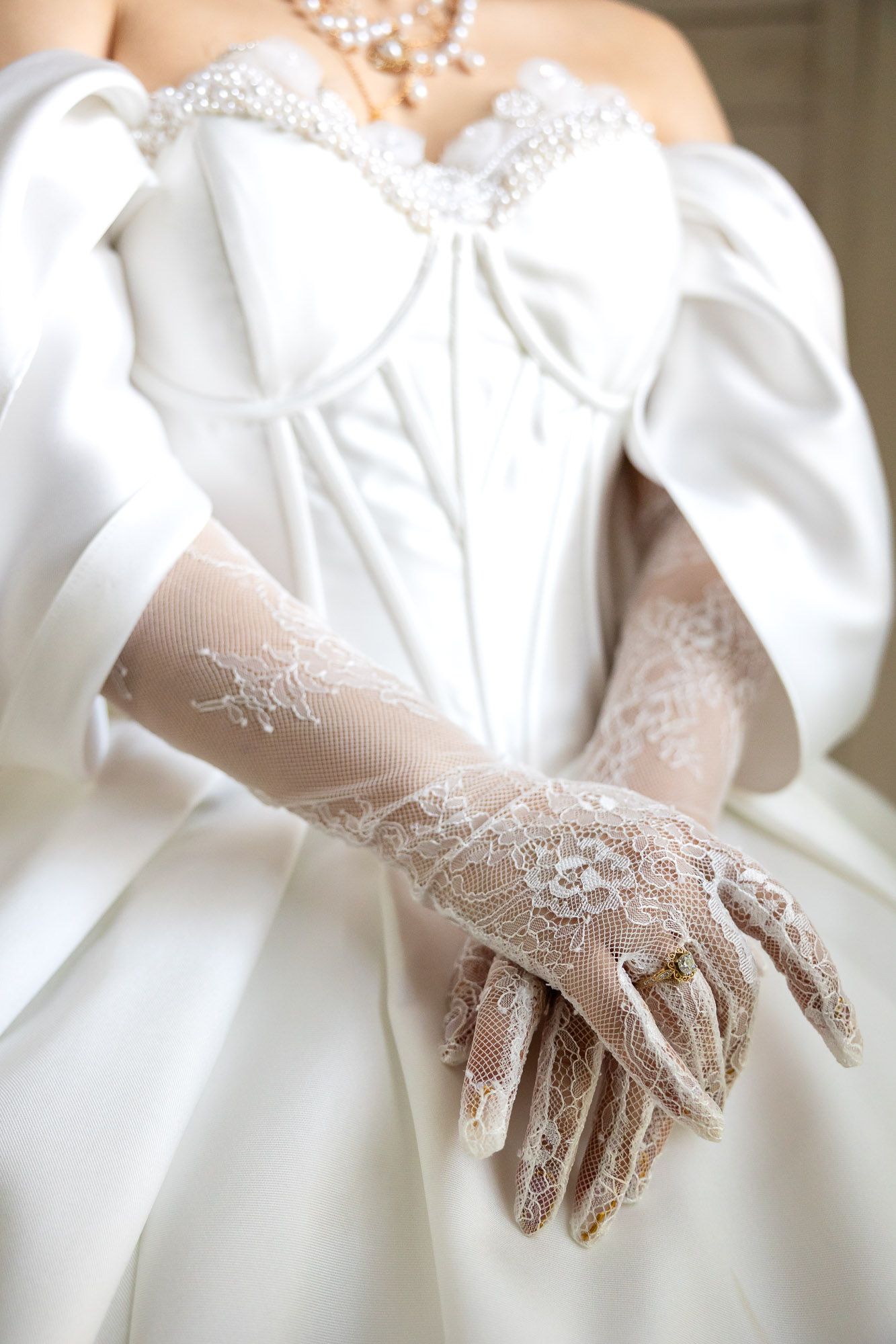 Bride in white satin corset gown wearing elegant lace gloves with pearl necklace on wedding day.