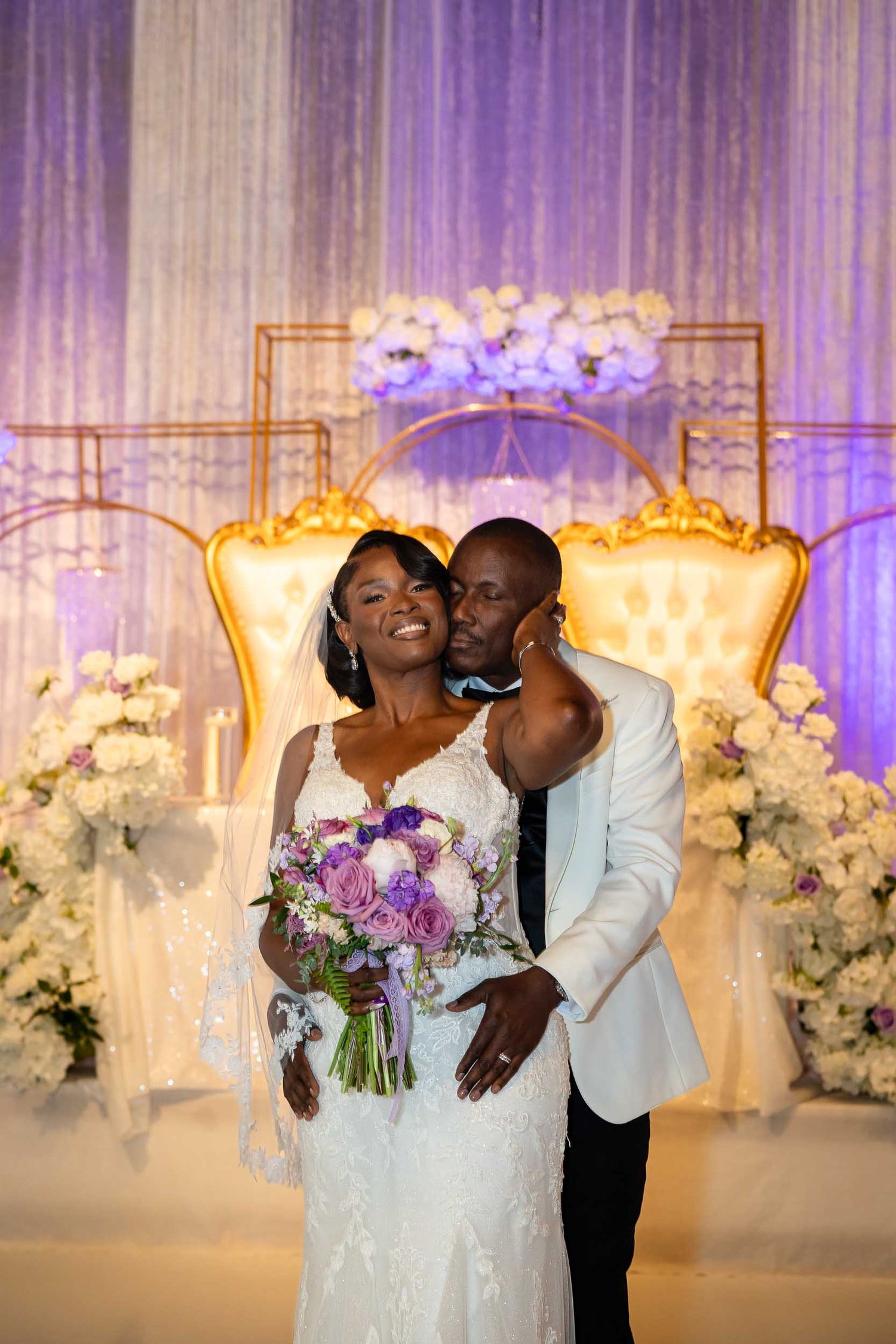 Bride and groom pose together at elegant wedding altar with purple draping and floral arrangements.