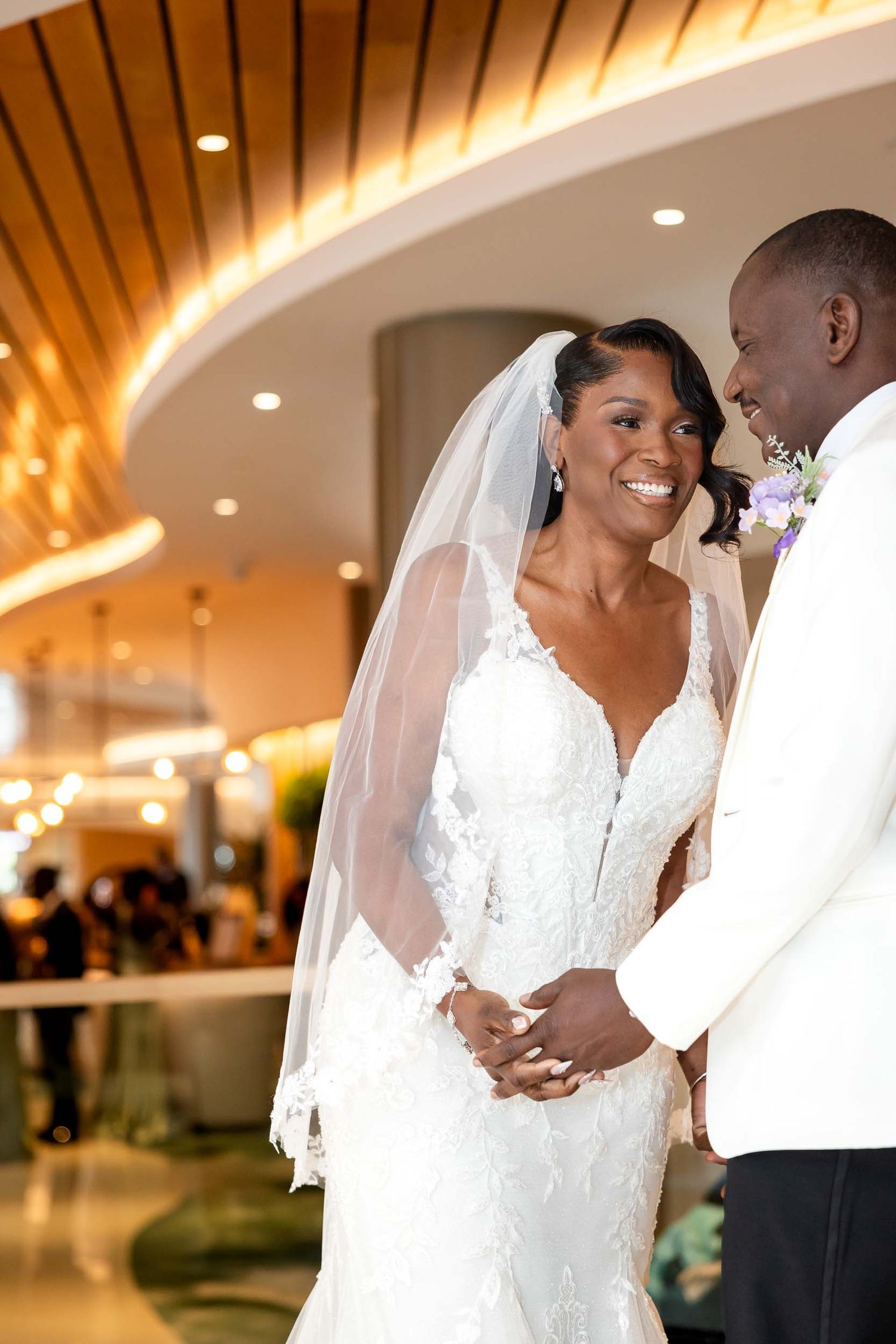 Smiling bride in lace gown and veil stands with groom in white tuxedo at elegant wedding venue.