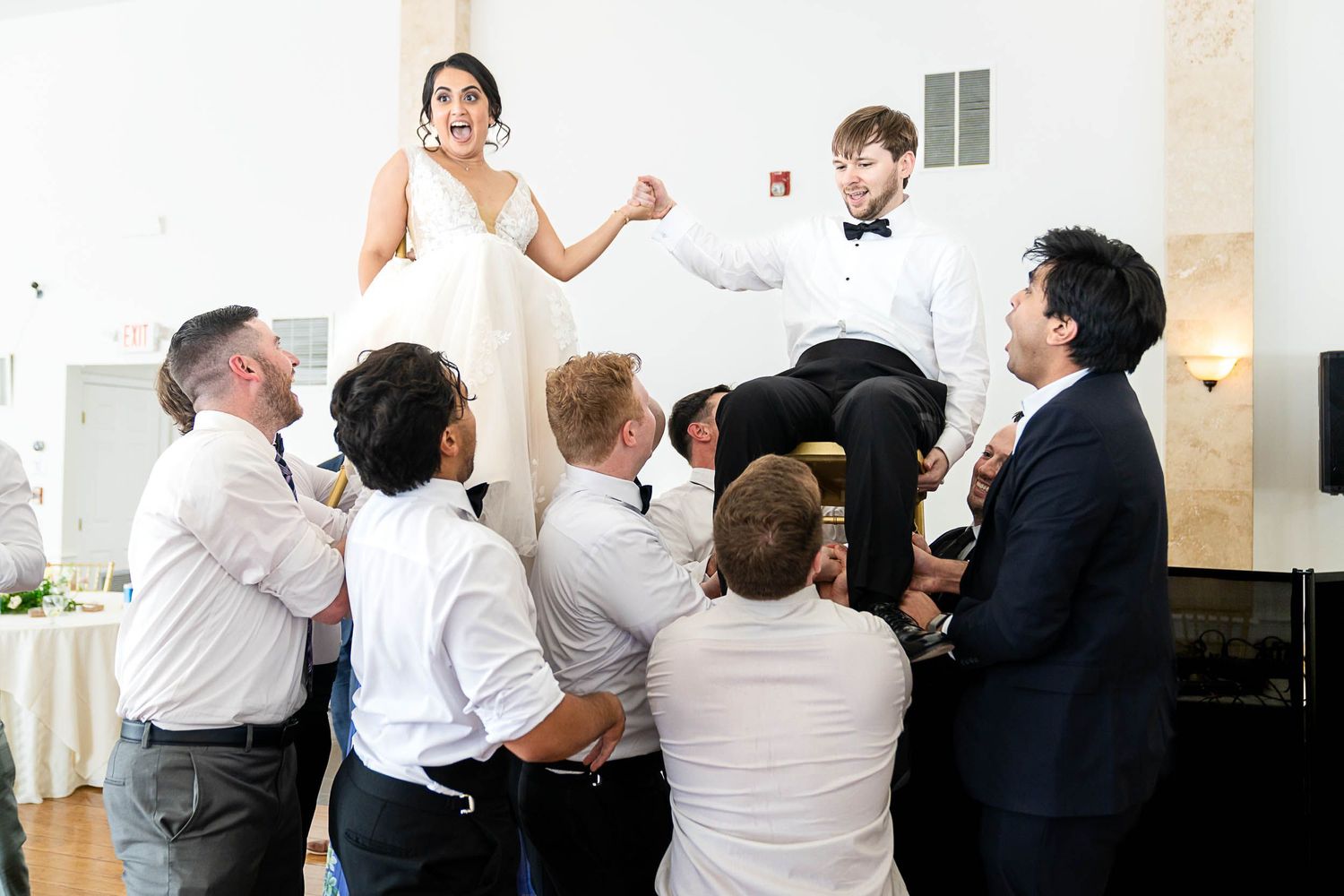 Wedding guests lifting bride and groom on chairs during festive reception celebration in elegant white venue.