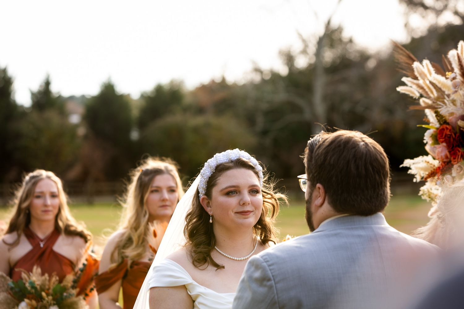 Bride smiling at groom during outdoor wedding ceremony with bridesmaids in rust dresses and pampas grass floral arrangements.