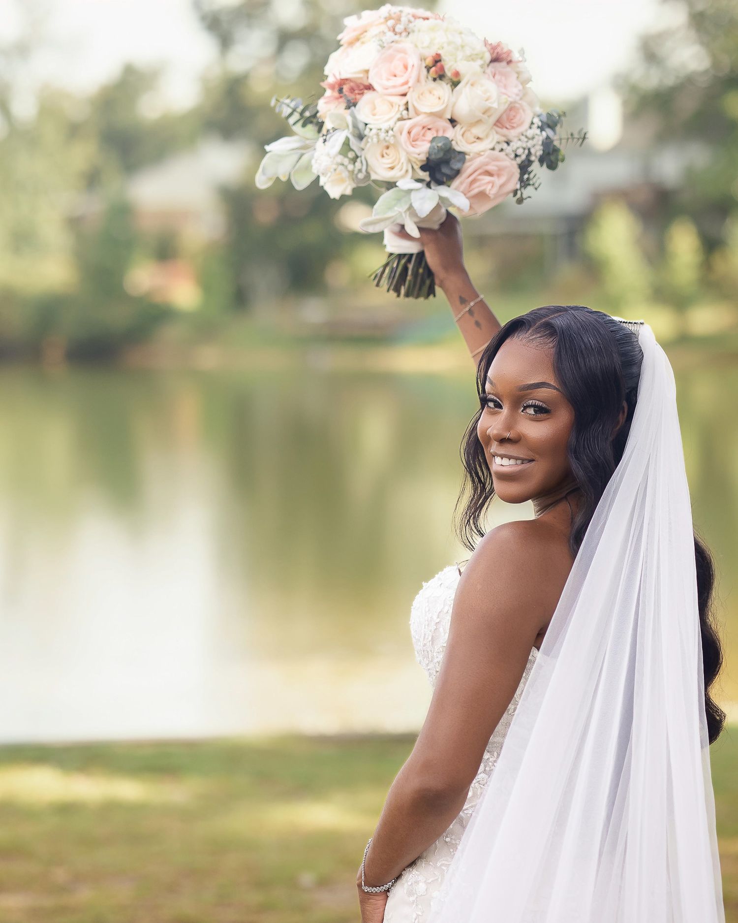 Smiling bride in white gown and veil holds bouquet aloft outdoors near a lake on her wedding day.