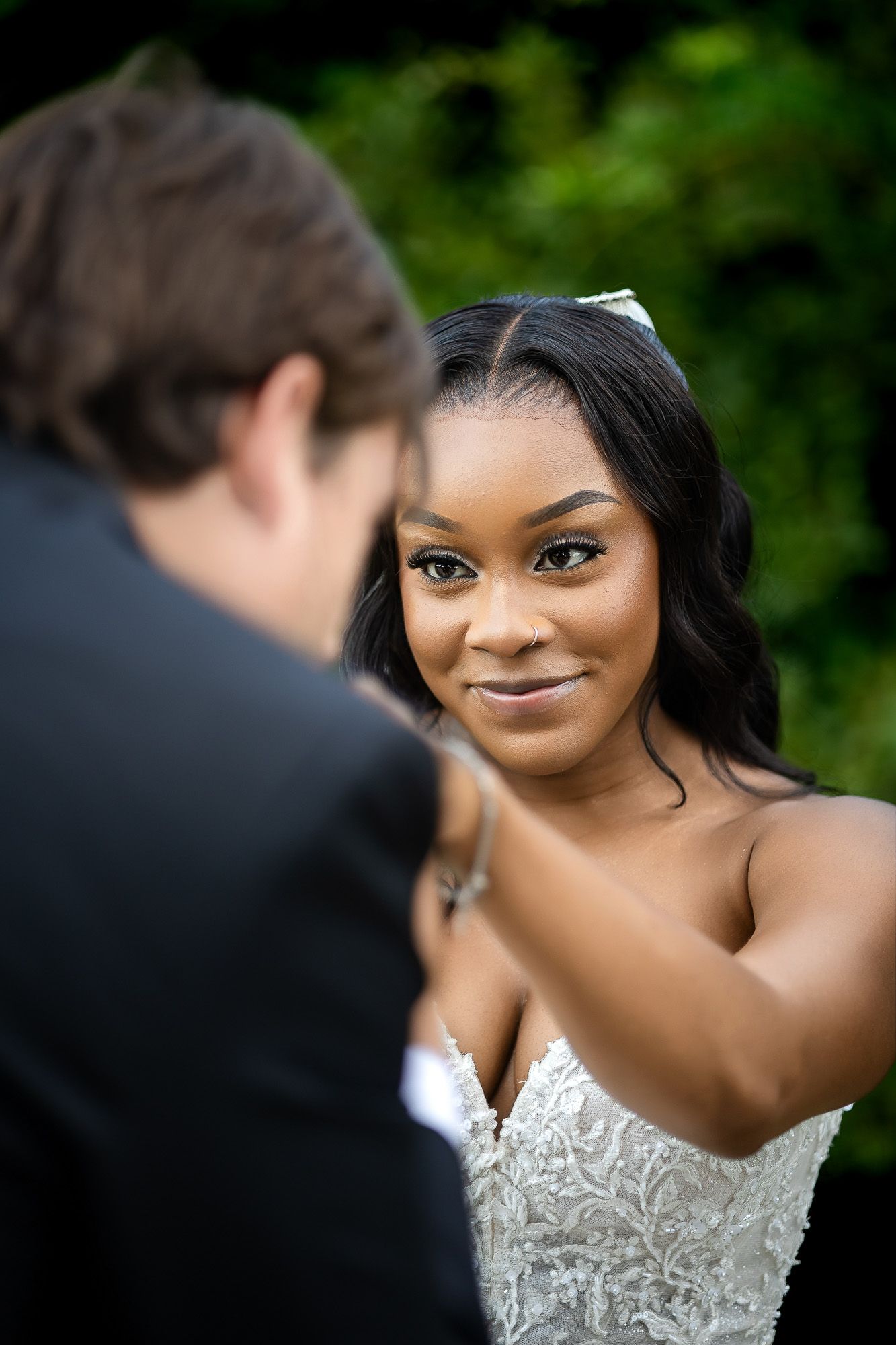 A bride in a lace gown smiles warmly at her groom during an outdoor wedding ceremony.