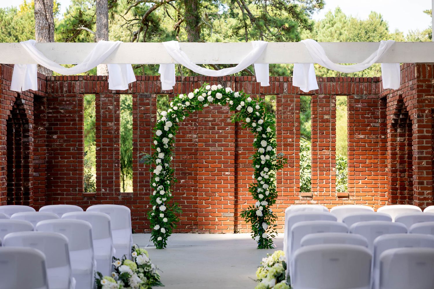 Elegant outdoor wedding ceremony setup with floral arch, white chairs, and brick wall backdrop surrounded by lush greenery.