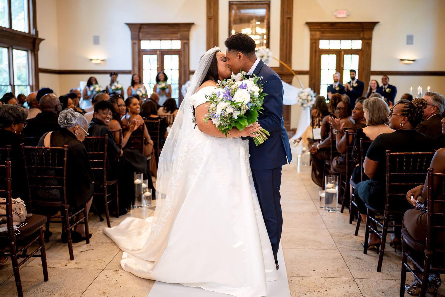 Bride and groom share a first kiss at wedding ceremony aisle, bride holding white and purple bouquet.