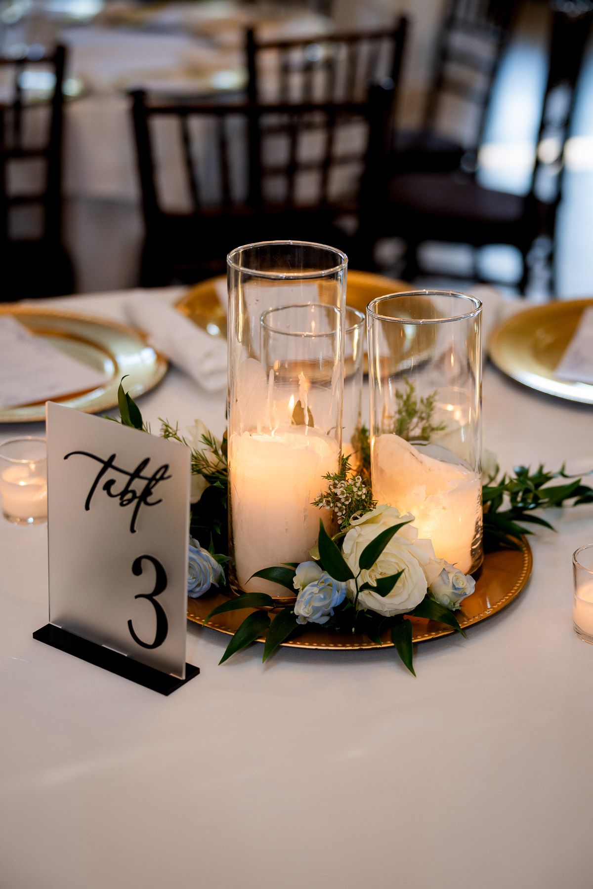 Elegant wedding table centerpiece with white candles, greenery, and flowers on a gold charger beside a Table 3 sign.