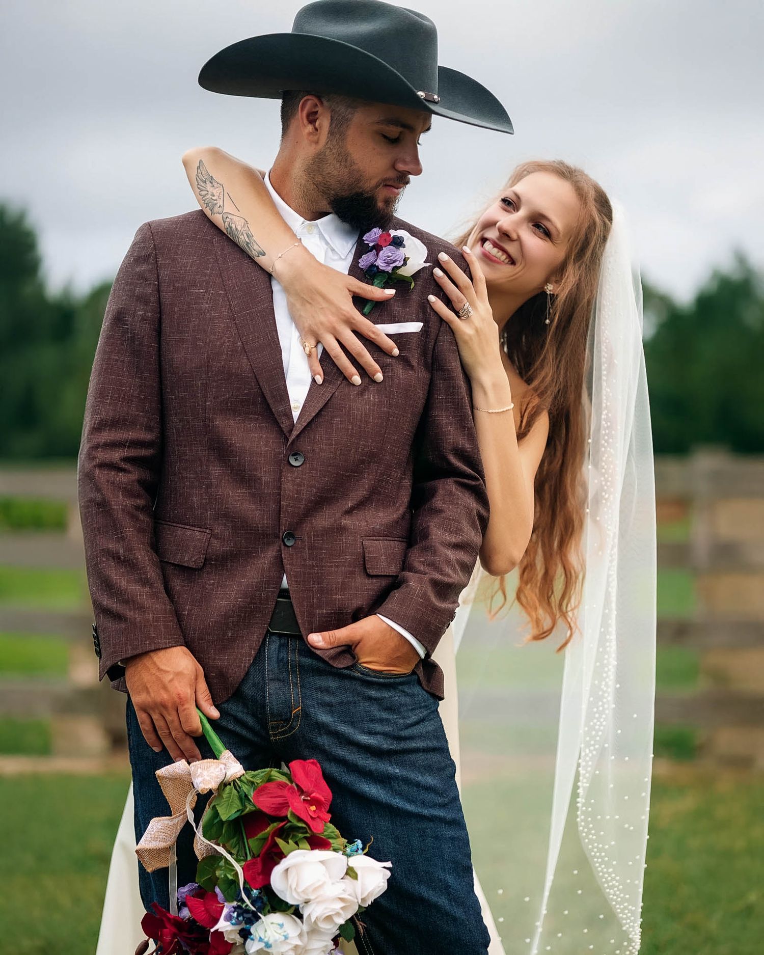 Couple in western wedding attire, groom in black cowboy hat and blazer, bride in veil holding bouquet, outdoor farm setting.