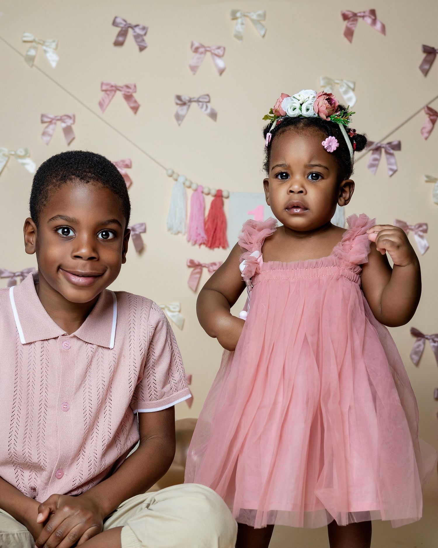 Two young children in pink outfits pose before a decorative bow backdrop, girl wearing floral crown.
