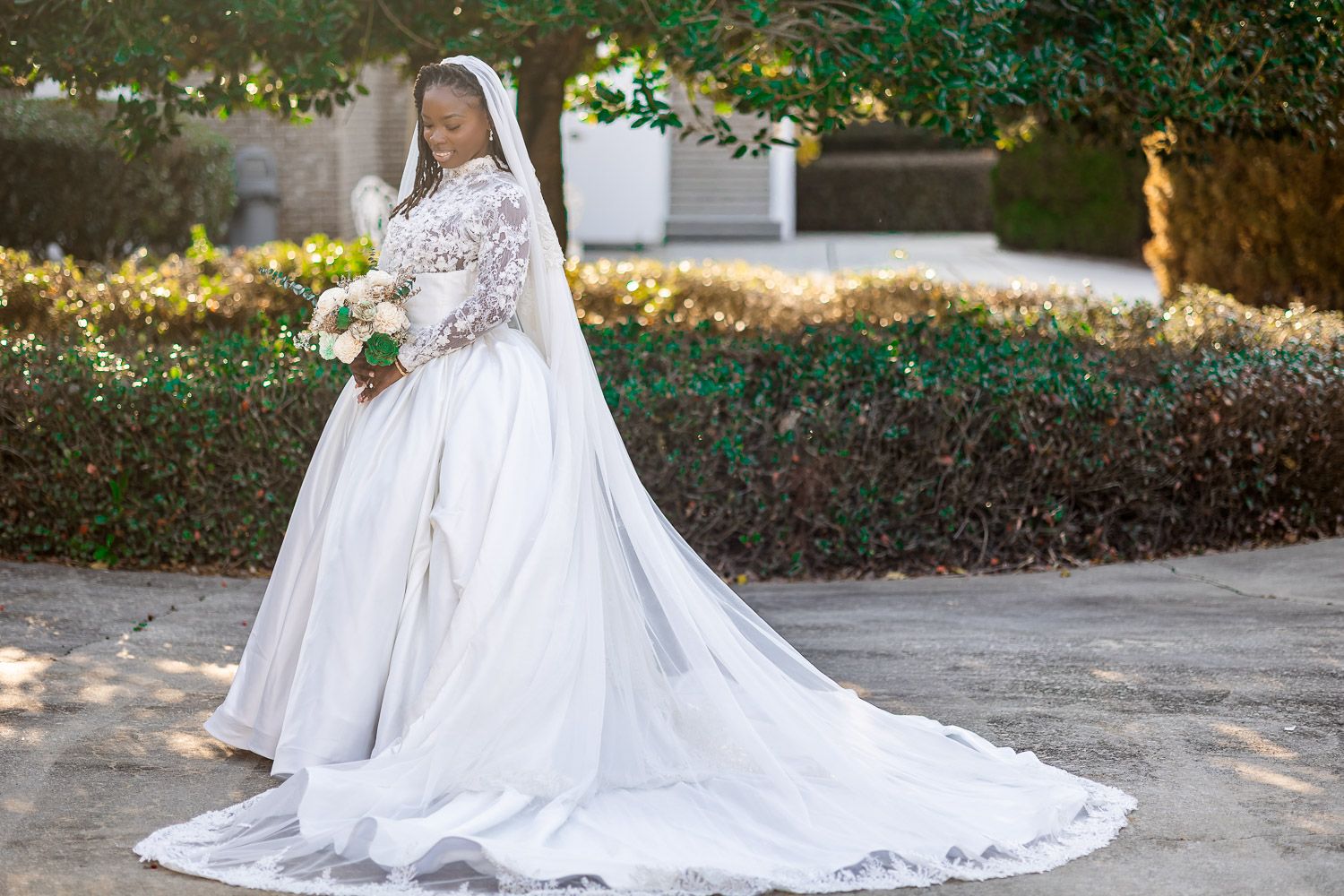 Bride in elegant long-sleeve white wedding gown with lace bodice and cathedral train holding bouquet outdoors.