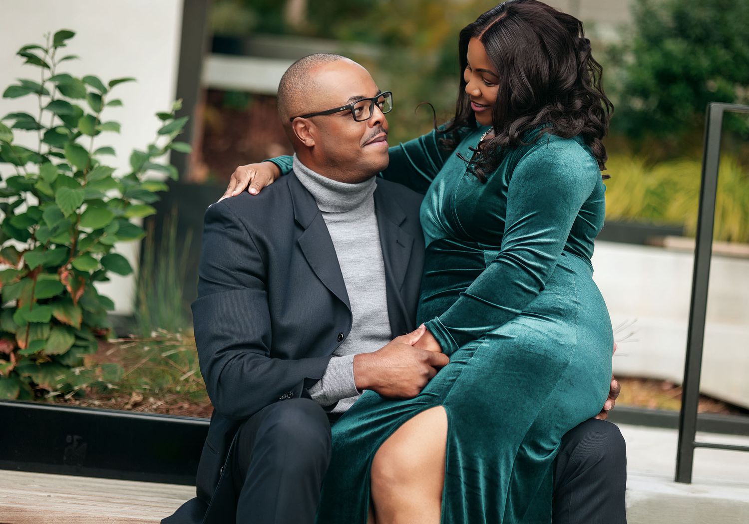 A couple shares an intimate moment outdoors, woman in teal velvet dress embracing a man in a gray turtleneck suit.