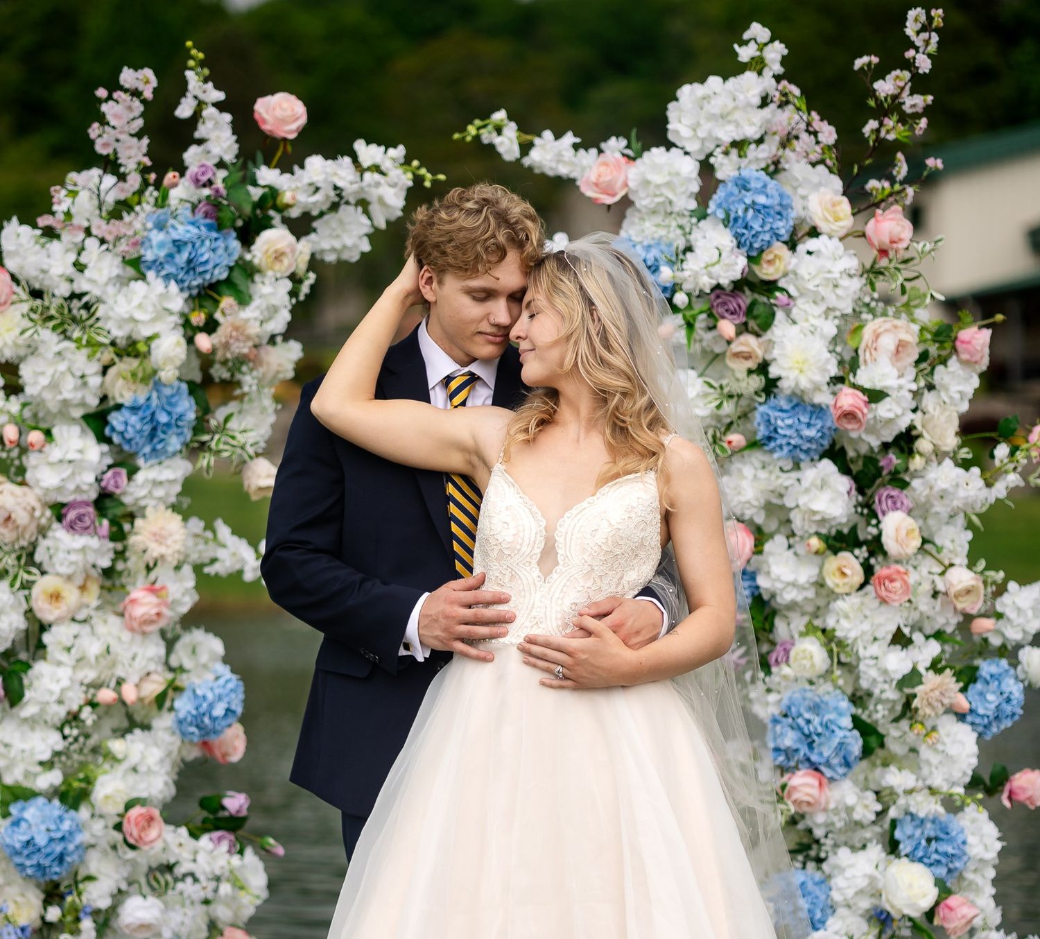 A bride and groom embrace under a floral arch adorned with blue hydrangeas and pink roses at an outdoor wedding ceremony.