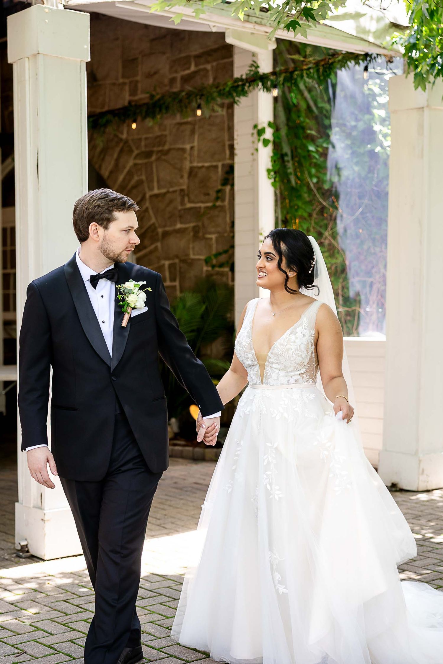 Bride in lace ballgown and groom in black tuxedo walk hand in hand outside a stone venue surrounded by greenery.