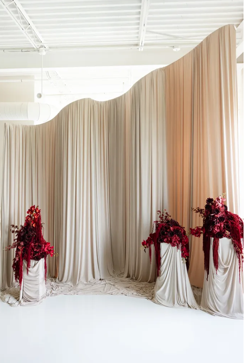 Elegant wedding backdrop with flowing ivory drapes and three white pedestals adorned with dramatic deep red floral arrangements.